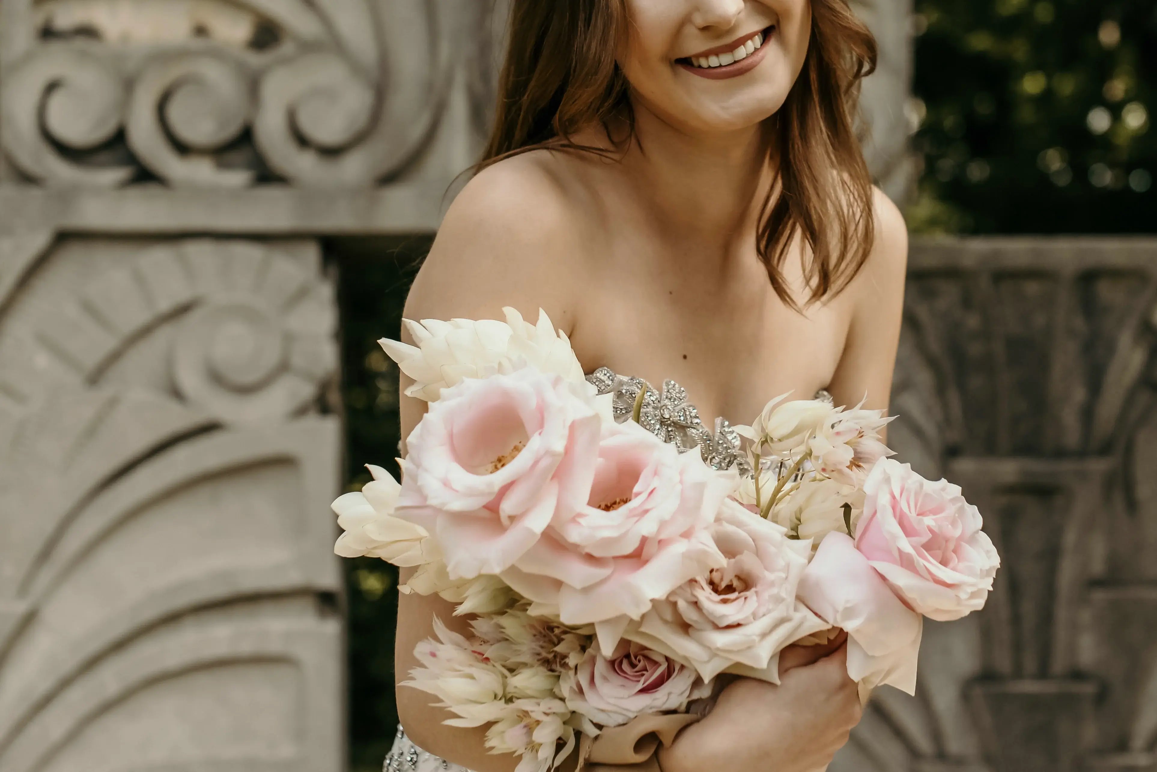 Woman holding a bouquet of pink and white flowers in front of an ornate stone wall.